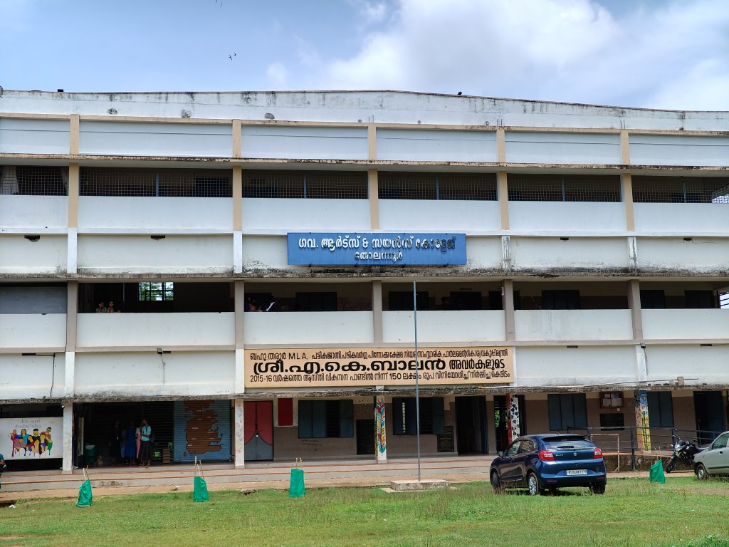 Front view of Government Arts and Science College, Tholanur, showing a three-story building with a blue sign, surrounded by greenery.