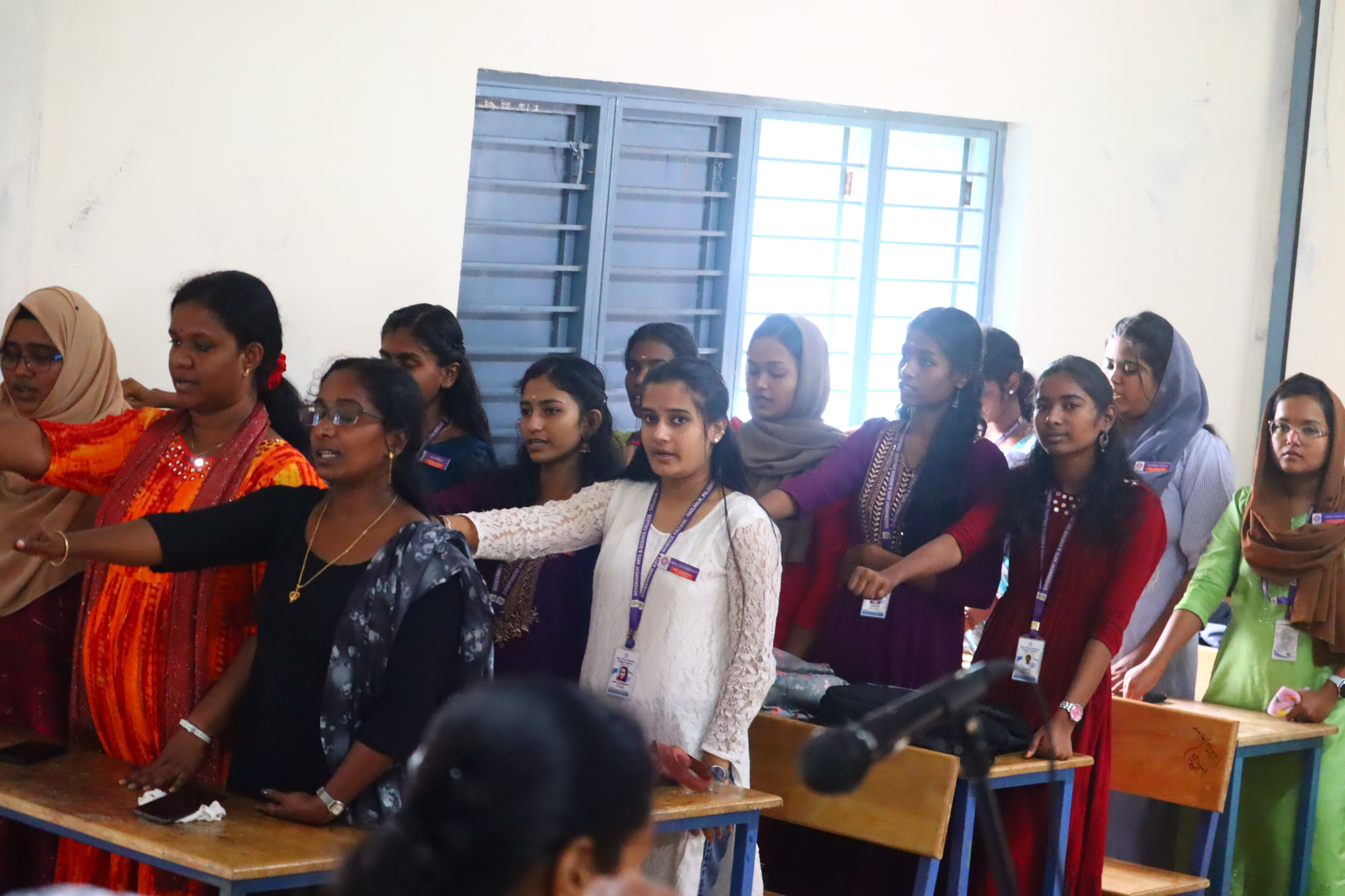 A group of female students standing in a classroom, with some raising their hands and others looking towards the front. The students are dressed in diverse traditional attire, with a range of colored outfits and accessories.