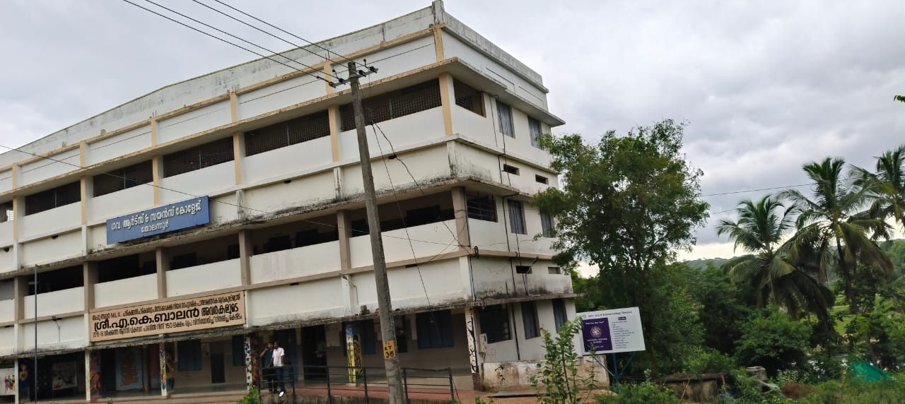 Exterior view of Government Arts and Science College, Tholanur, showcasing a three-story building with a signboard, surrounded by greenery and cloudy skies.