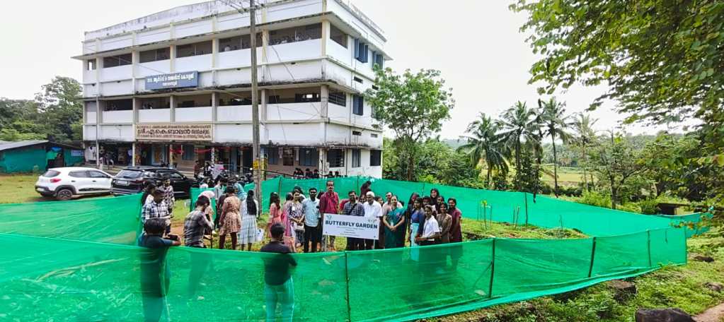 Group of people standing behind a green mesh fence at the Butterfly Garden, with a building and trees in the background.
