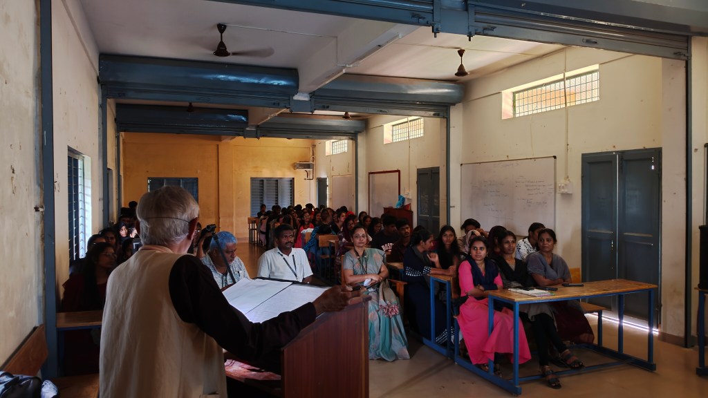 A classroom setting with a speaker addressing an audience. The room contains students seated at desks, with the speaker positioned at the front. There are windows and walls painted in warm colors, and the atmosphere suggests a learning environment.