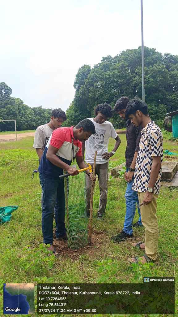 A group of six individuals gathered in a green field, examining a basket containing plants. They are engaged in discussion, with trees in the background providing a natural setting.