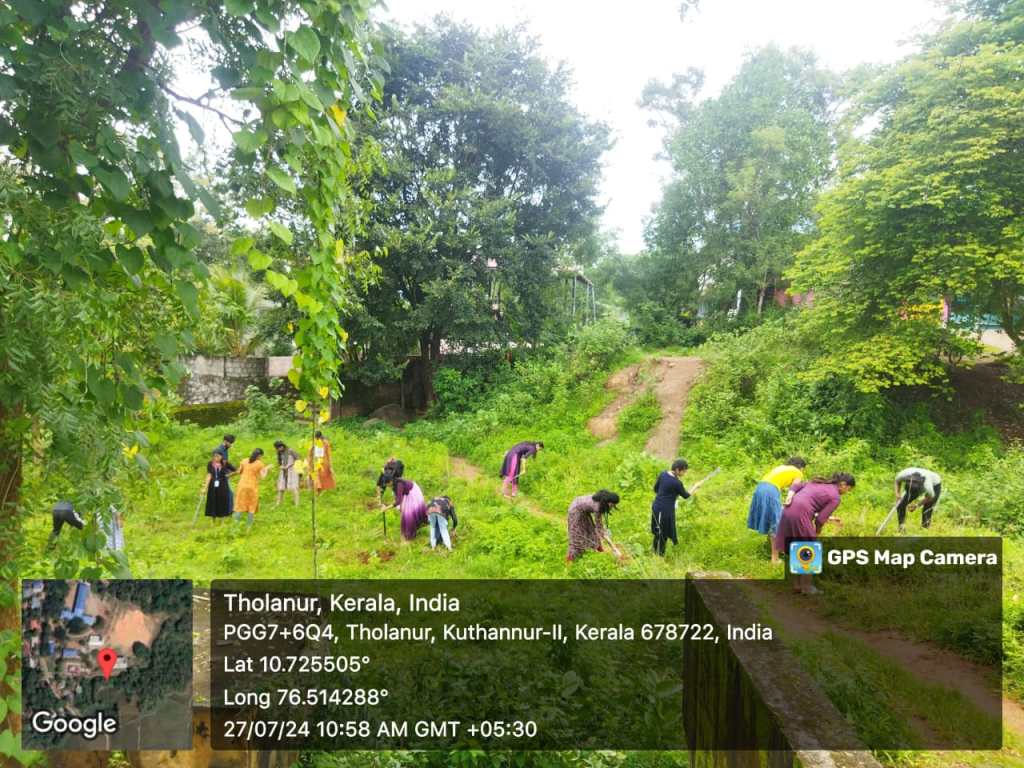 People working in a lush green field in Tholanur, Kerala, India, with trees in the background.