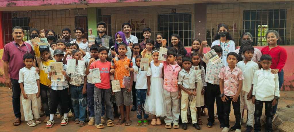 Group photo of children and adults holding up papers, standing outside a building with a pink wall, with some individuals wearing masks.