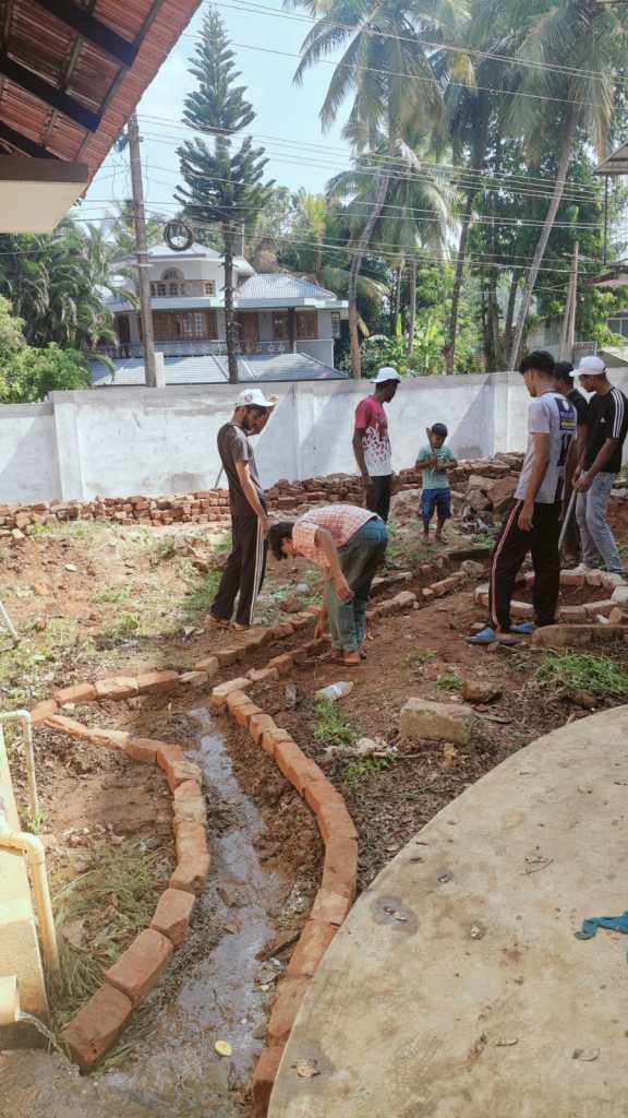 A group of people working outdoors in a garden, arranging bricks along a path next to a water stream, with palm trees and buildings in the background.