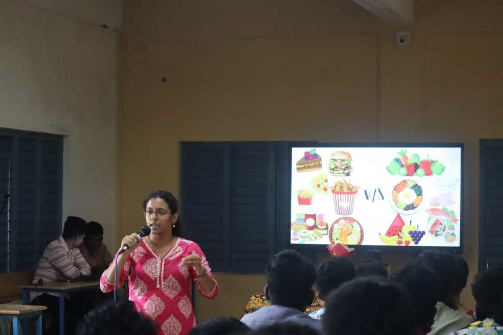 A speaker in a pink outfit presents a program on health, with food imagery on a screen behind them. Audience members are engaged in the session.